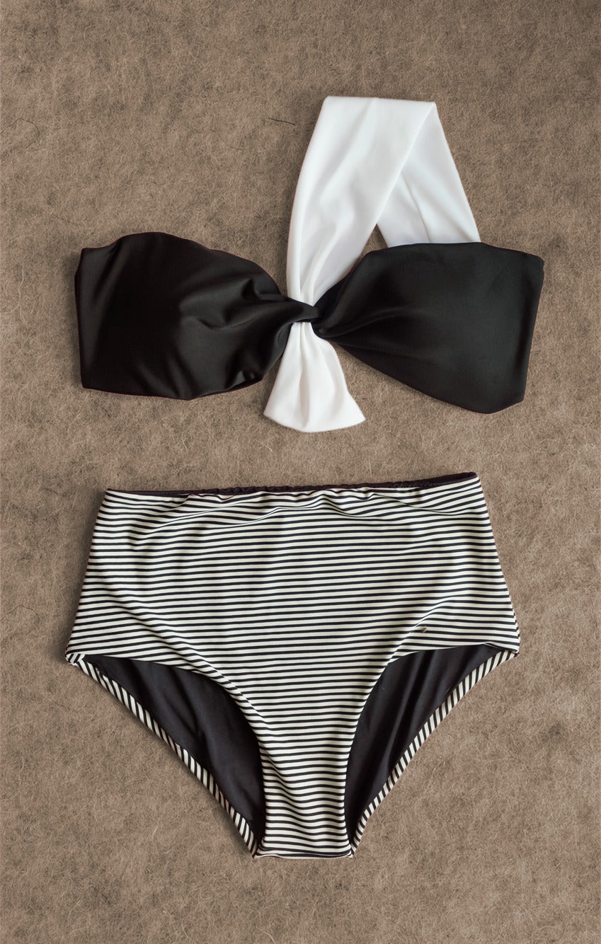 Luxurious two-tone striped bikini with a black bandeau top featuring a white knot, and coordinating high-waisted bottoms, from the Yacht Luxury Collection, presented on a textured background.