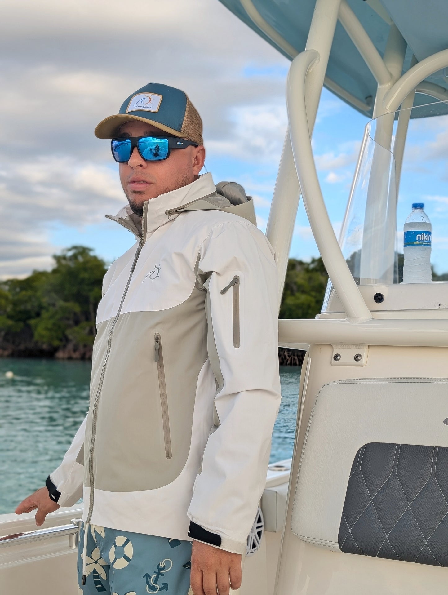 Man wearing waterproof adventure fishing jacket on a boat, facing forward, with tropical coastline in the background.