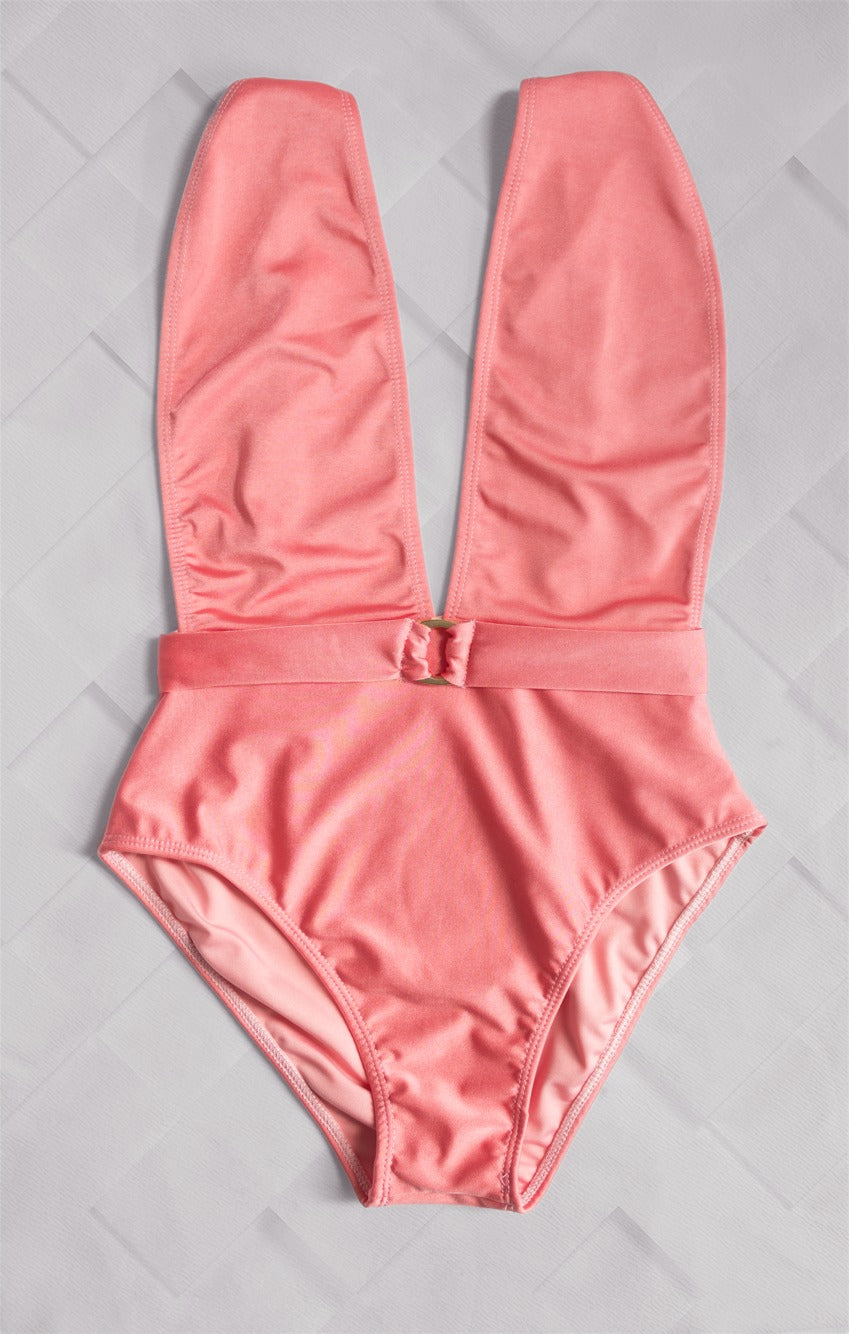 Luxury coral-colored one-piece swimsuit with a plunging neckline and belt detail from the Yacht Luxury Collection, presented against a geometric-patterned backdrop.