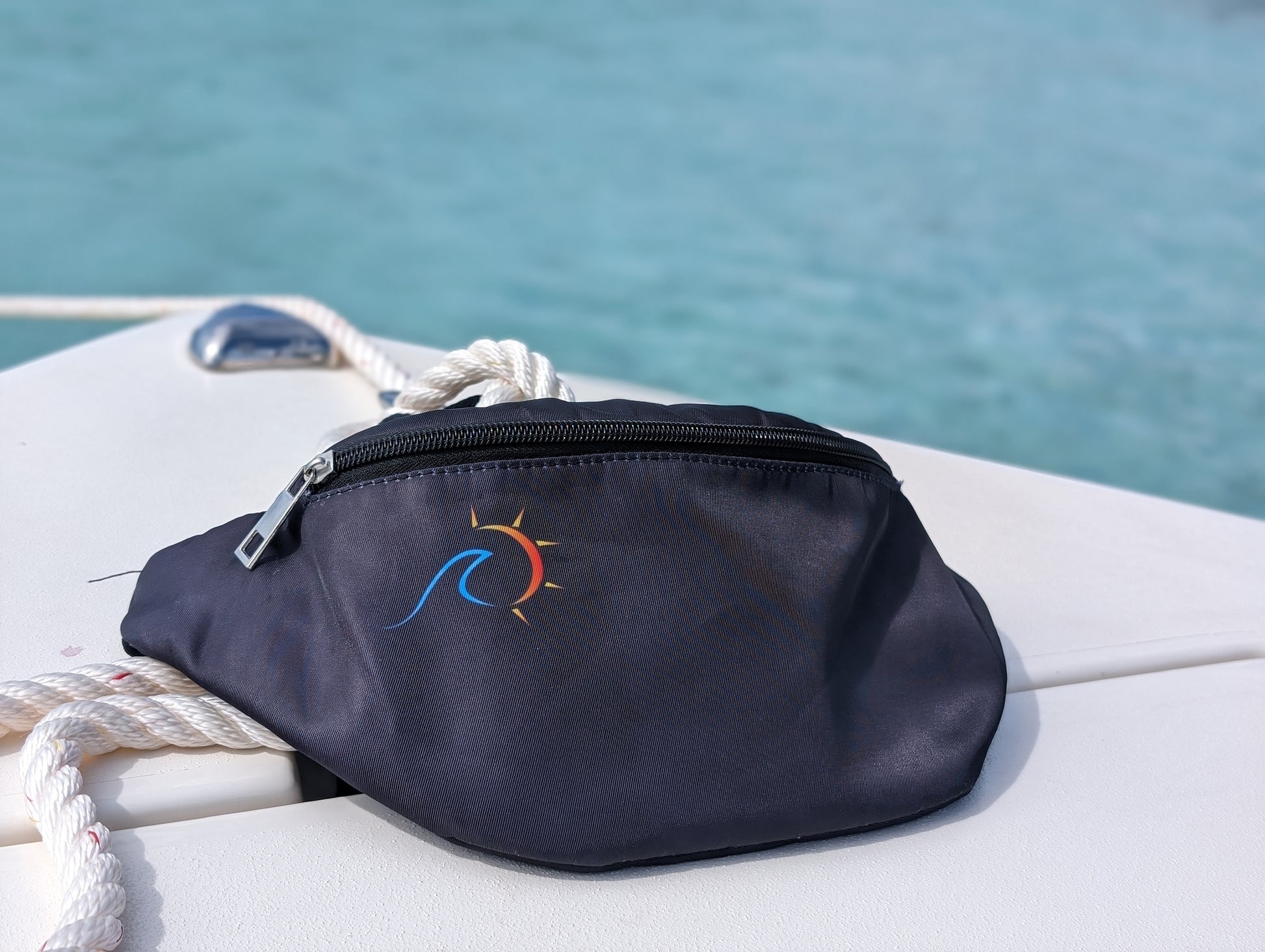 Black fanny pack with sublimated logo lying on a white surface with a rope coiled beside it.