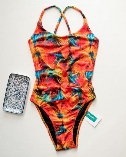 One-piece swimsuit laid flat with a vibrant abstract pattern and cross-back straps, paired with a decorative phone case and brand tag on a white background.