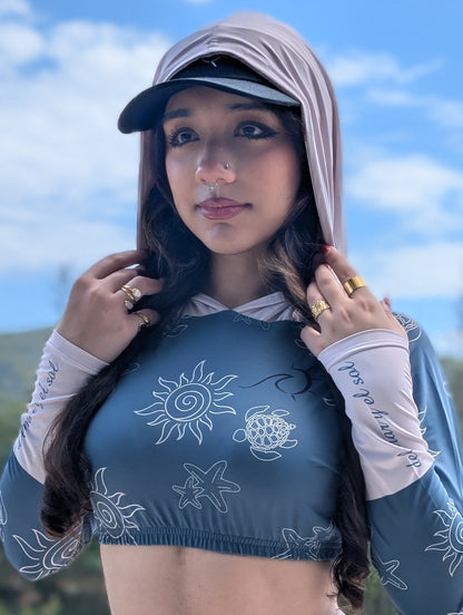 Woman wearing a blue deep turquoise crop rash guard with a sand color hoodie and del mar y el sol text on both sleeves 