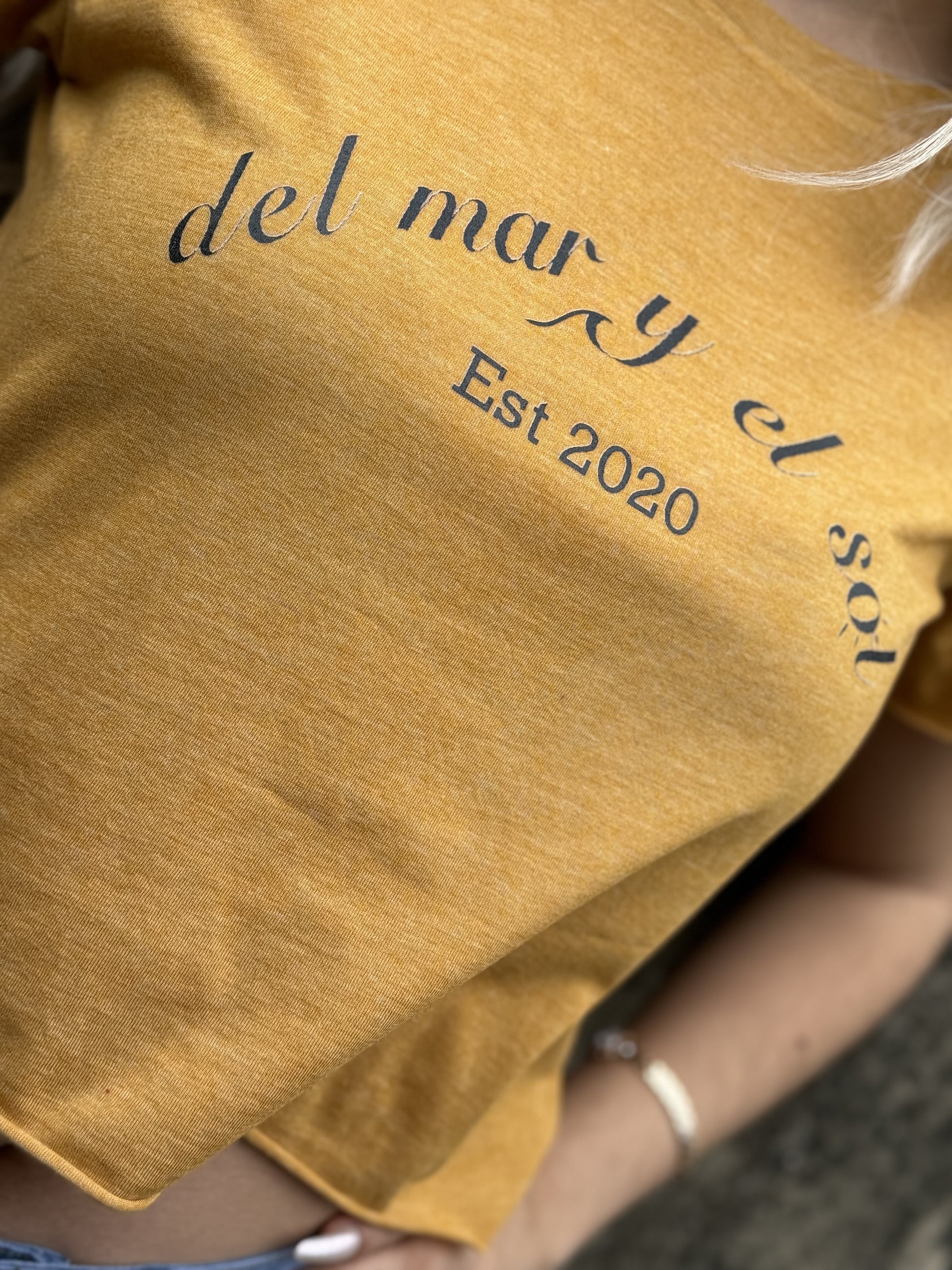 A person wearing a mustard cropped top with 'DEL MAR Y EL SOL' text on it, indicating the brand name and the phrase 'EST 2020'.
