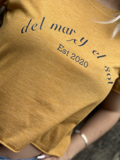 A person wearing a mustard cropped top with 'DEL MAR Y EL SOL' text on it, indicating the brand name and the phrase 'EST 2020'.