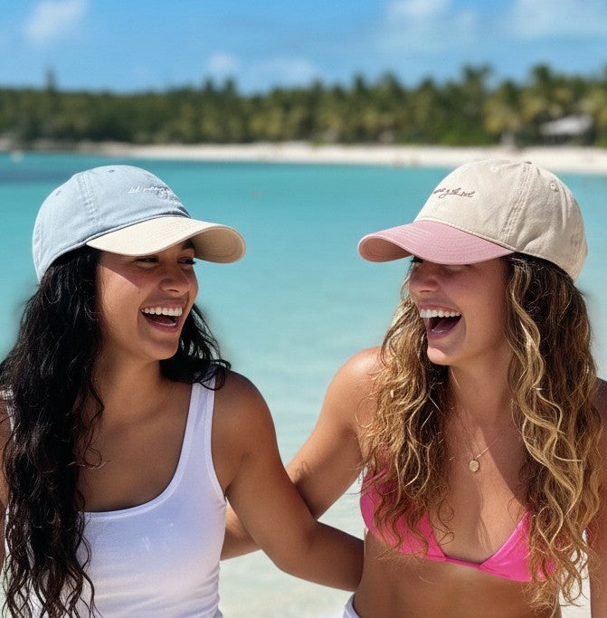 Two women laughing on a beach with del mar y el sol signature adjustable beach cap.
