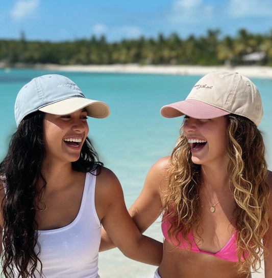 Two women laughing on a beach with del mar y el sol signature adjustable beach cap.