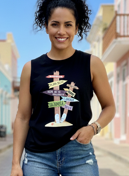 Woman wearing a black sleeveless tank shirt with a graphic of a direction sign with different name of beaches in Puerto Rico with denim shorts standing on a colorful street.