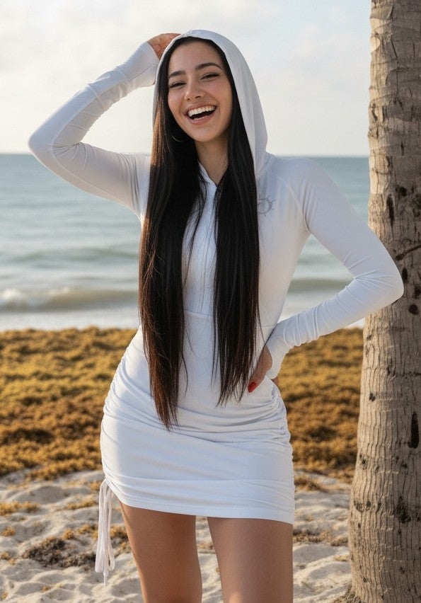 Woman in a white hooded dress with adjustable length and a front zipper, standing by a palm tree on a beach.