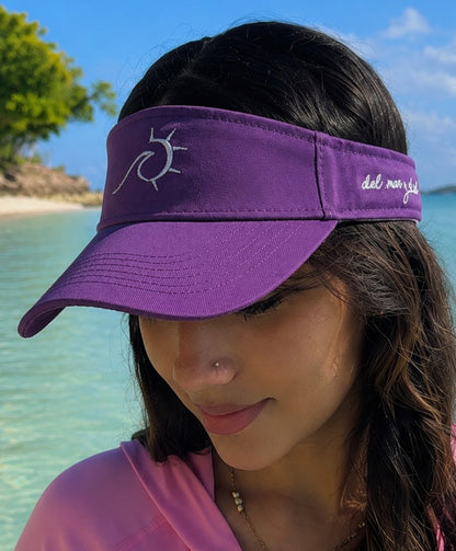 A woman wearing a purple visor with a white sun logo and the text 'del mar y el sol' on the side, photographed outdoors at a beach with turquoise water and blue sky in the background.