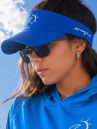 A woman wearing a bright blue long-sleeve cropped sun hoodie with white script text down the sleeve, a matching blue visor, and dark swim bottoms, sitting on the edge of a boat