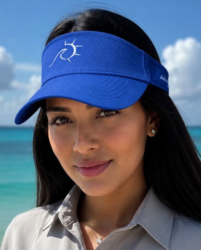 A woman wearing a royal blue visor with a white sun logo, photographed outdoors against a beach and blue sky background.
