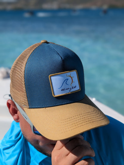 A person wearing a blue and beige breathable mesh trucker hat with a curved visor with a del Mar y el Sol badge on it.
