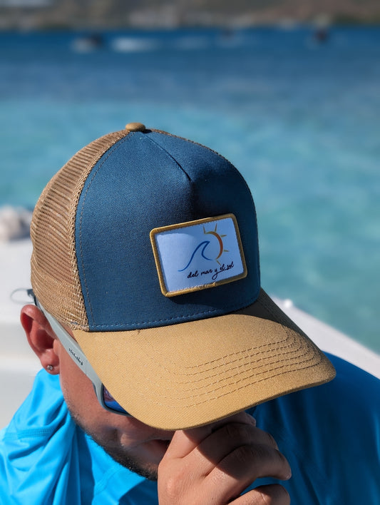 A person wearing a blue and beige breathable mesh trucker hat with a curved visor with a del Mar y el Sol badge on it.