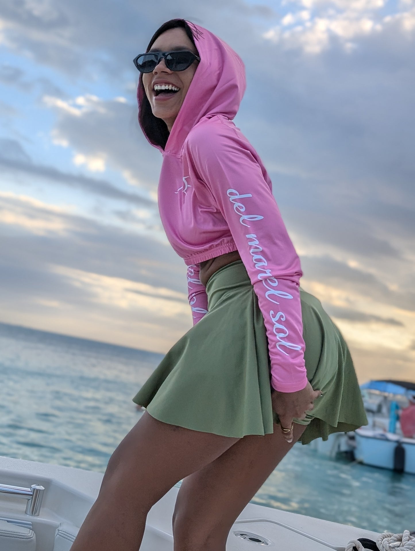 A woman wearing a pink cropped rash guard with hoodie, standing on a boat with the ocean in the background. The rash guard has text on the sleeve.