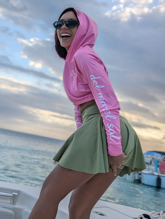 A woman wearing a pink cropped rash guard with hoodie, standing on a boat with the ocean in the background. The rash guard has text on the sleeve.